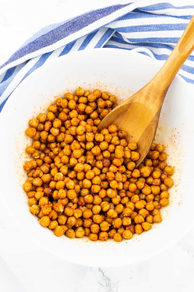 roasted chickpeas in a white bowl with a wooden spoon on a marble counter top with a blue dishtowel in the background