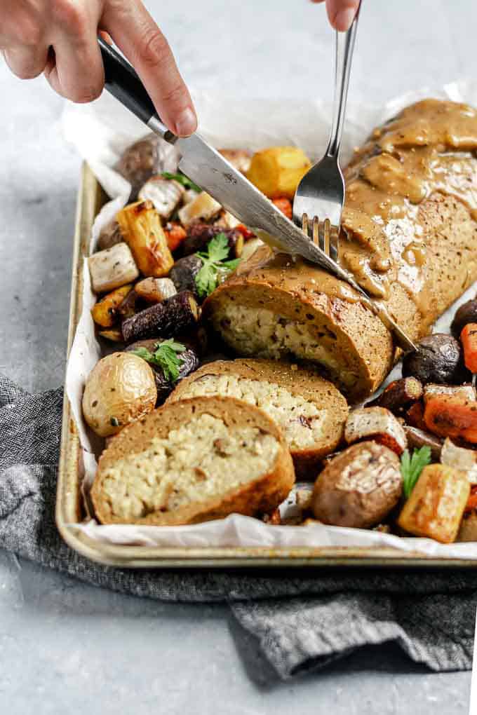 a seitan roast on a baking tray with baked vegetables and potatoes is being cut into slices with a fork and a knife 
