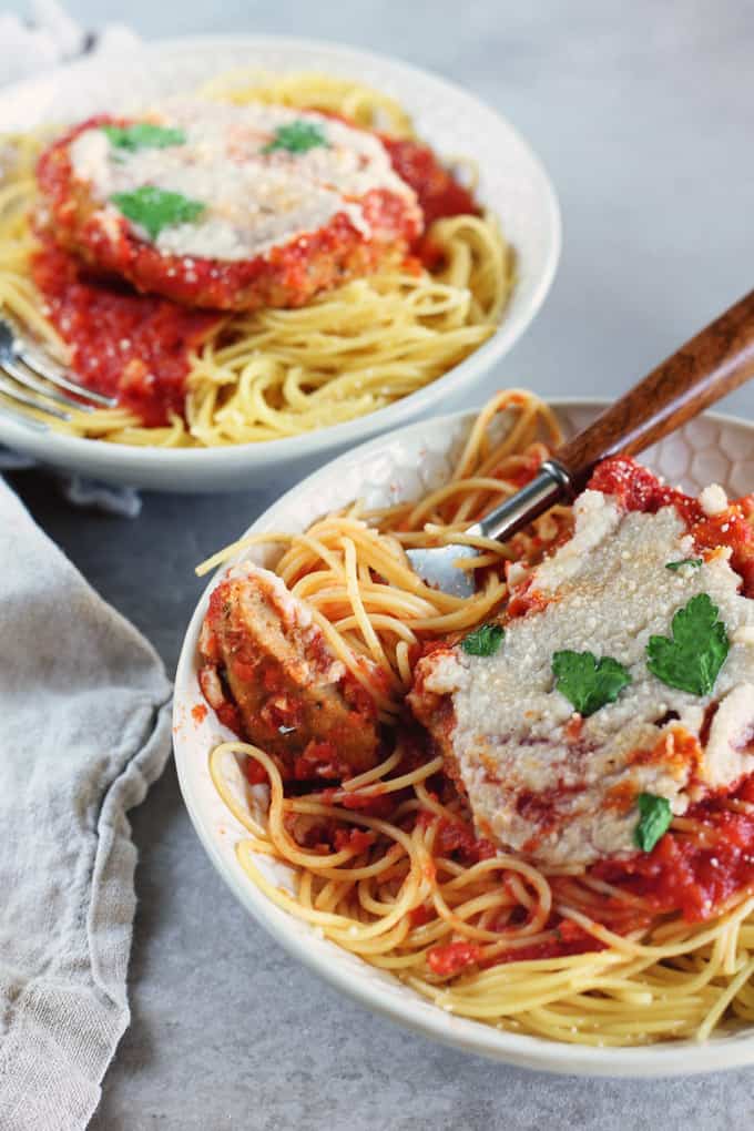 two bowls with spaghetti, tomato sauce, and vegan seitan Parmesan with a fork and a grey table cloth on the side 