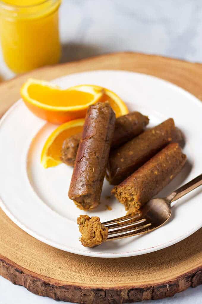 three vegan breakfast sausages on a white plate with a fork and three orange slices on the side and a glass of orange juice in the background 