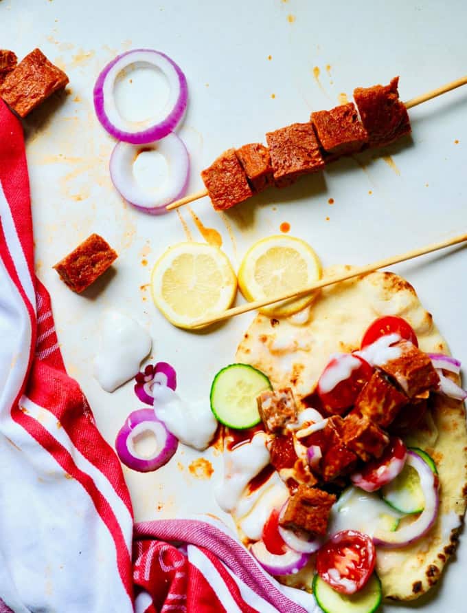 Greek Vegan Seitan Gyros on skewers on a white counter top with red onion rings, cucumber, lemon, and a filled pita on the side 