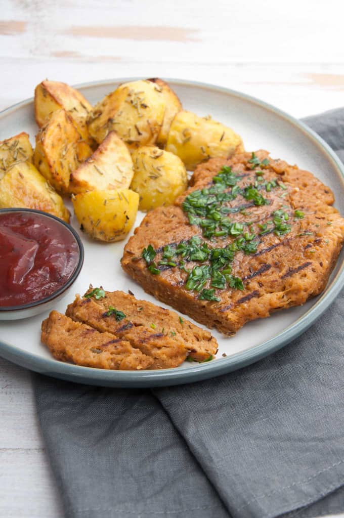 a vegan seitan steak on a plate with potato wedges and a small bowl of ketchup on the side 