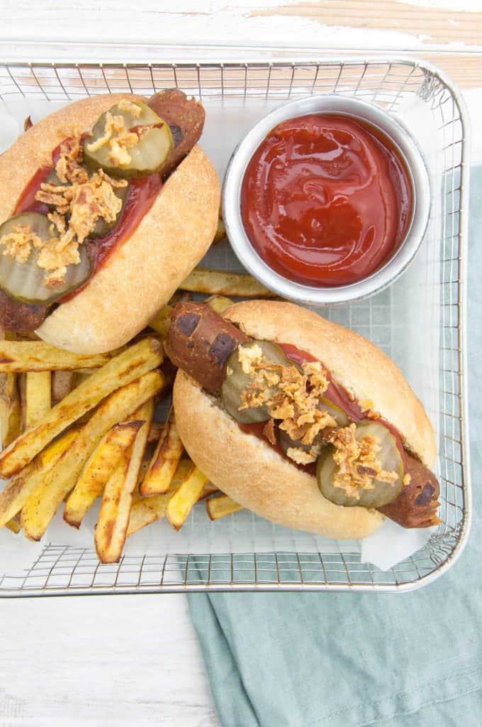 two vegan hot dogs in a basket with fries and a little bowl of ketchup on a white wooden board 