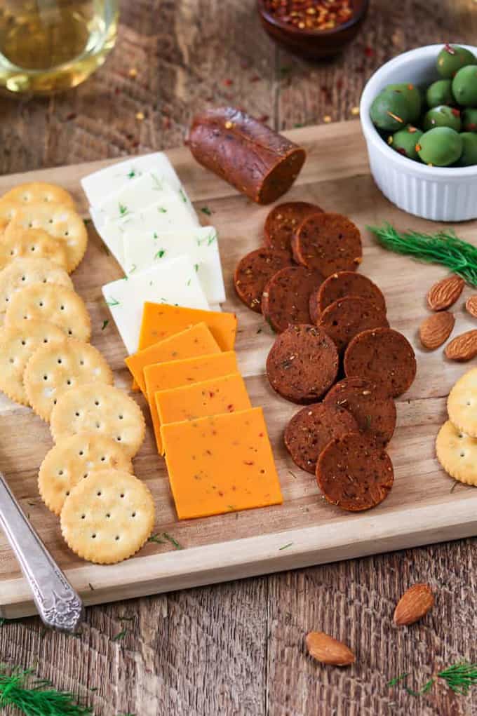 vegan pepperoni, vegan cheese, and crackers on a wooden cutting board with a small bowl of olives and almonds and a glass of wine in the background