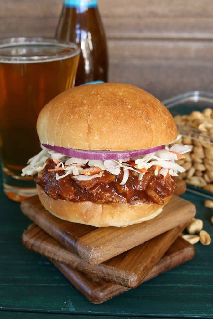 a sausage seitan sandwich on a stack of three small wooden boards with a bottle and a glass of beer in the background 