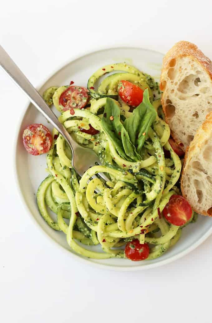 zucchini noodles with pesto and cherry tomatoes on a white plate with two slices of bread on the side on a white counter top 