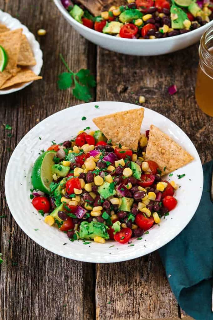 Avocado Black Bean Corn Salad in a white bowl with corn chips on a wooden board with another bowl of corn salad in the background 