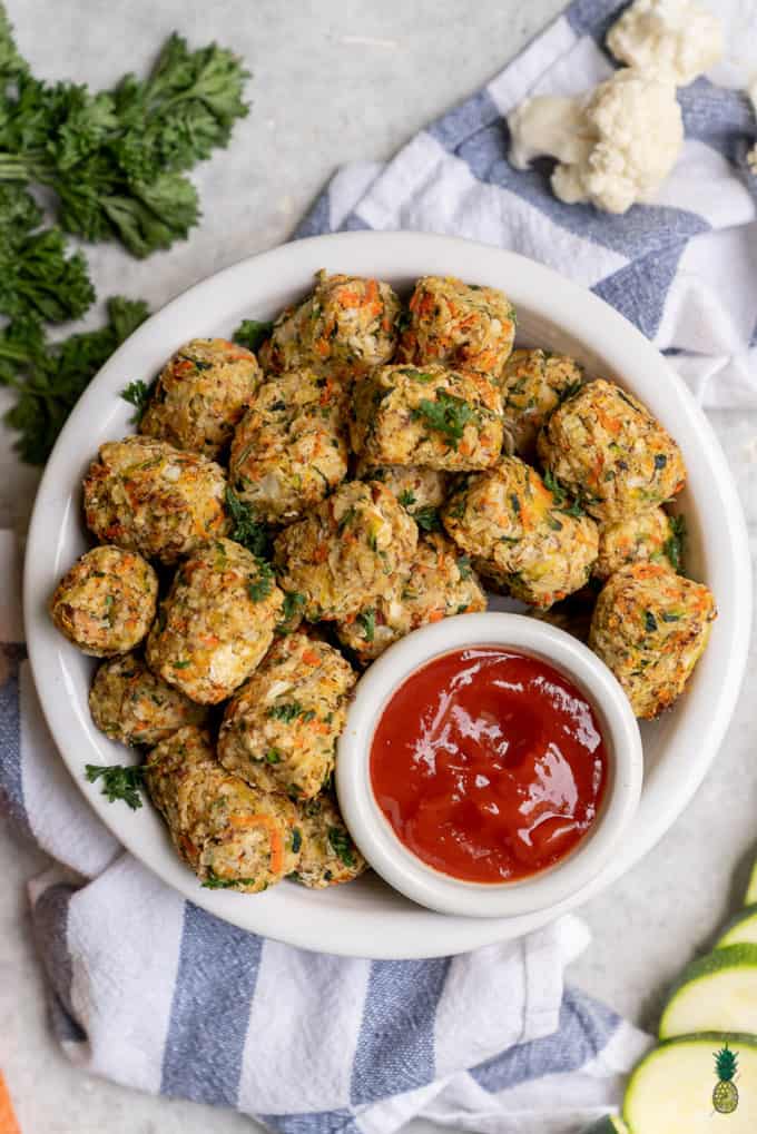 baked veggie tater tots in a white bowl with a small bowl of ketchup on a striped white and blue table cloth 