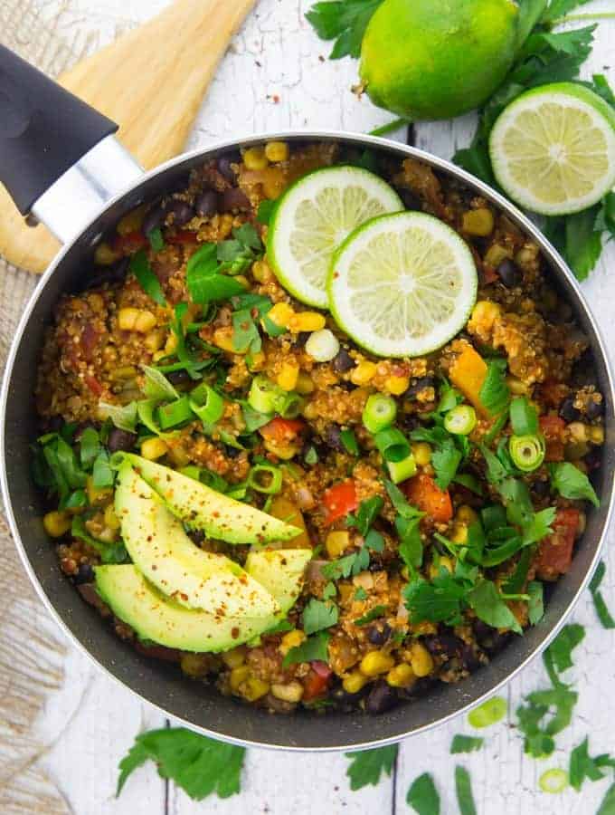 a pot of Mexican quinoa with avocado slices, lime halves, and chopped parsley on top on a white wooden board 