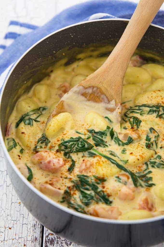a pot with vegan gnocchi in a creamy sauce with spinach and tomatoes with a wooden spoon 