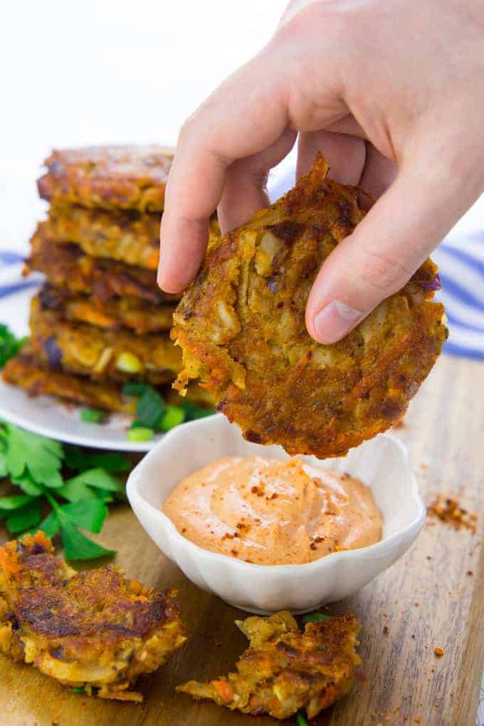 a hand dipping a potato fritter into a small bowl of vegan sriracha mayonnaise with more fritters on a plate in the background