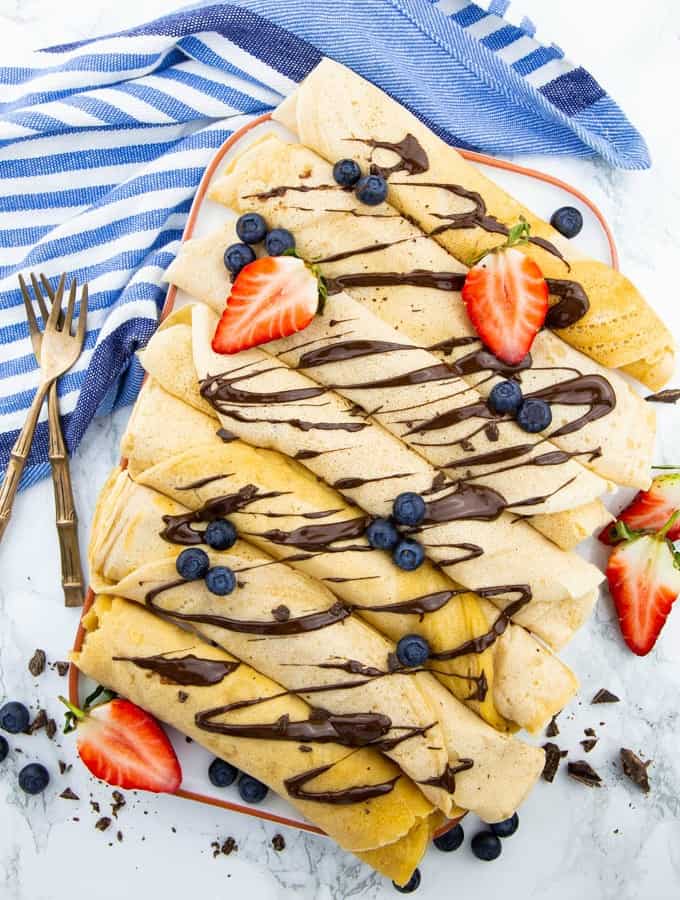 seven vegan crepes on a white plate on a marble counter top with chocolate sauce, strawberries, and blueberries on top