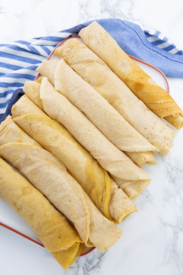 seven rolled up vegan crepes on a white plate on a marble counter top
