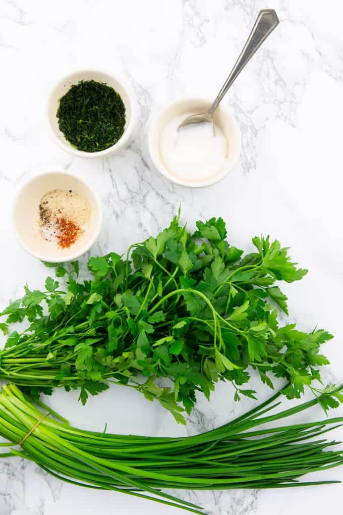 fresh chives, parsley, and three little bowls of vegan mayonnaise, chopped dill, and spices on a marble counter top (ingredients for vegan ranch) 