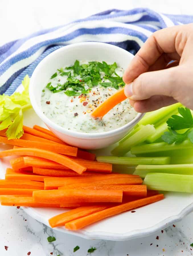a hand dipping a carrot stick in a small bowl of vegan ranch on a plate with carrot and celery sticks 