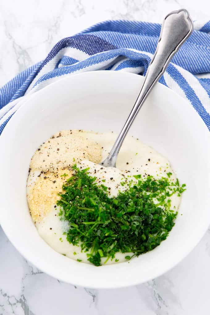 vegan mayonnaise, dill, parsley, chives, onion, and garlic powder in a white bowl with a spoon on a marble counter top (preparation of vegan ranch) 