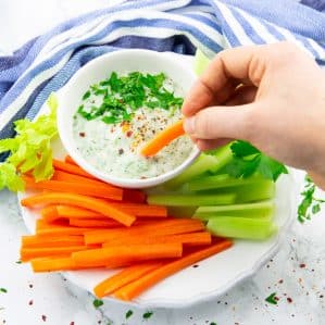 a hand dipping a carrot stick in a small bowl of vegan ranch on a plate with carrot and celery sticks