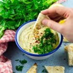 Baba Ganoush in a white and blue bowl on a blue counter top with parsley in the background and a hand dipping a pita into the Baba Ganoush