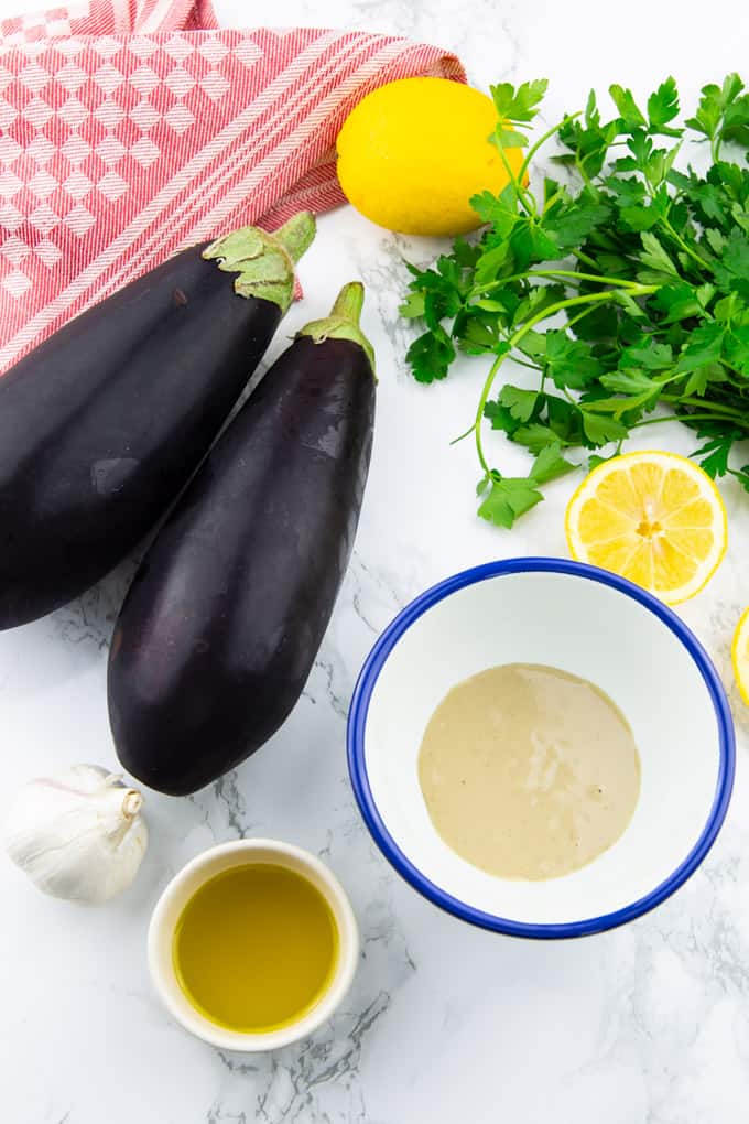 two eggplants, a small bowl of olive oil, a bowl of tahini, garlic, parsley, and two lemons on a marble counter top 