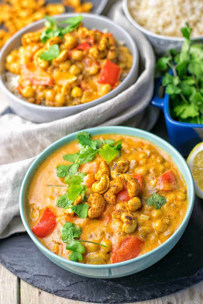 two bowls of Lentil Chickpea Yellow Curry on a wooden counter top with a bowl of rice in the background 