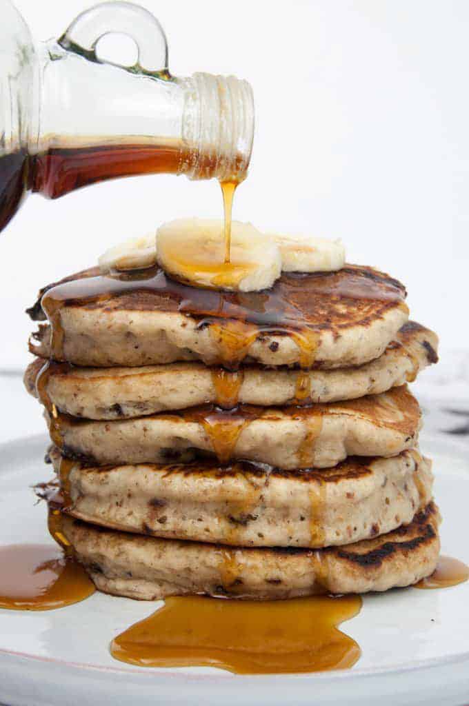 a stack of vegan banana pancakes on a white plate with maple syrup being poured over the pancakes 