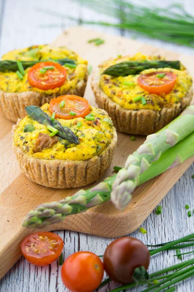 three vegan mini quiches on a wooden board with green asparagus and cherry tomatoes on the side 