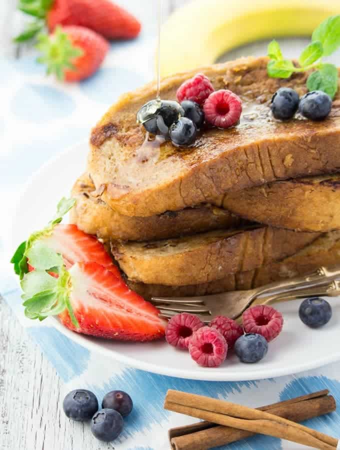 four slices of vegan French toast on a white plate with raspberries, blueberries, and strawberries on the side 