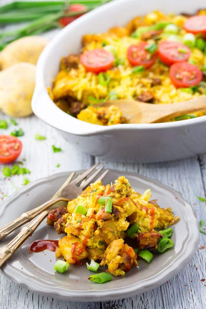 vegan breakfast casserole with potatoes, tofu scramble, and tempeh on a grey plate with two forks with a casserole dish in the background