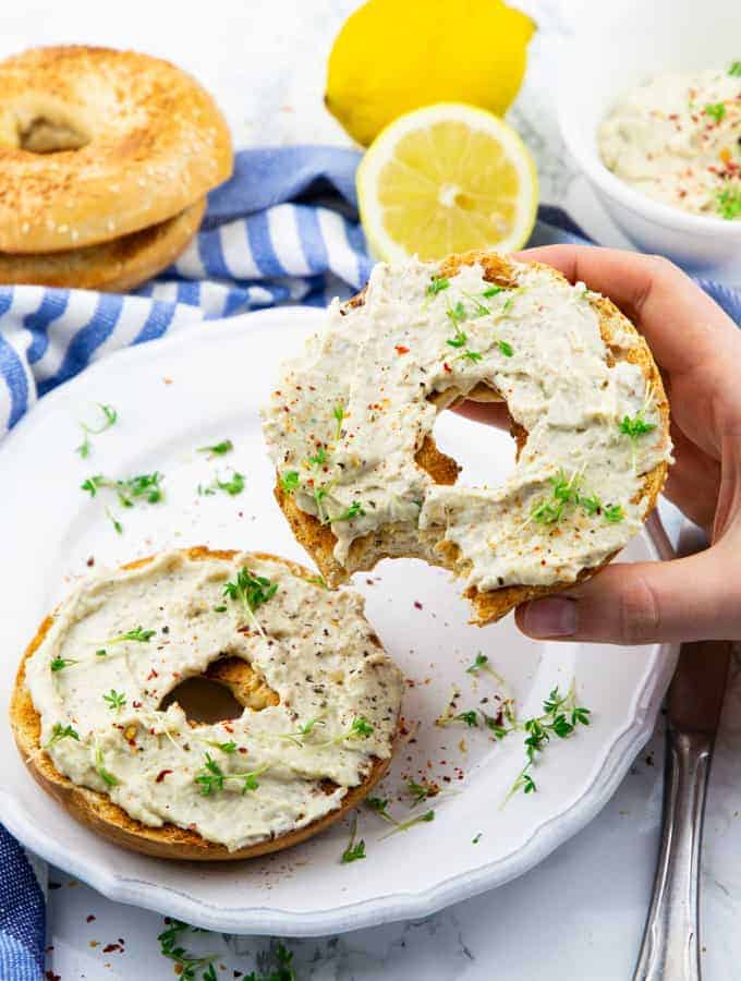 a hand holding a bagel with vegan cream cheese over a white plate with another bagel and a blue dishcloth