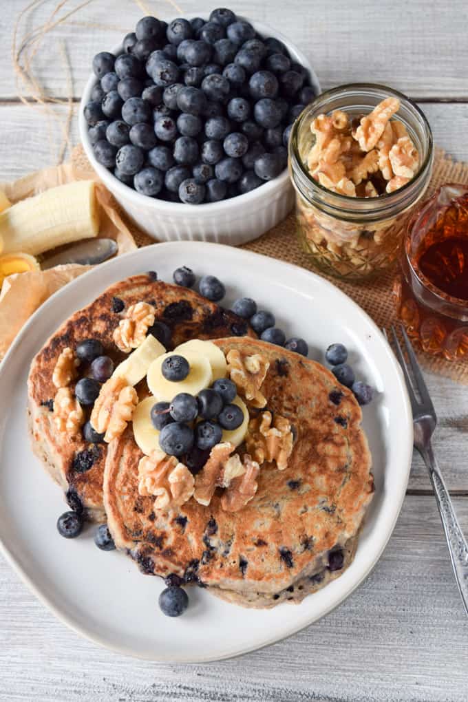 two vegan blueberry pancakes on a white plate on a white wooden counter top with blueberries and walnuts in the background 