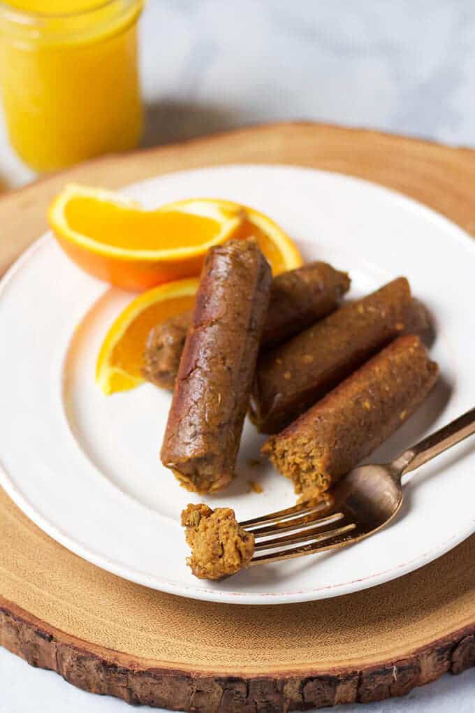 three vegan maple breakfast sausages on a white plate with a fork on a marble counter top with a glas of orange juice in the background 