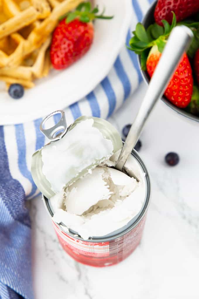 an opened can of coconut milk with a spoon on a marble counter top with a plate of waffles and strawberries in the background