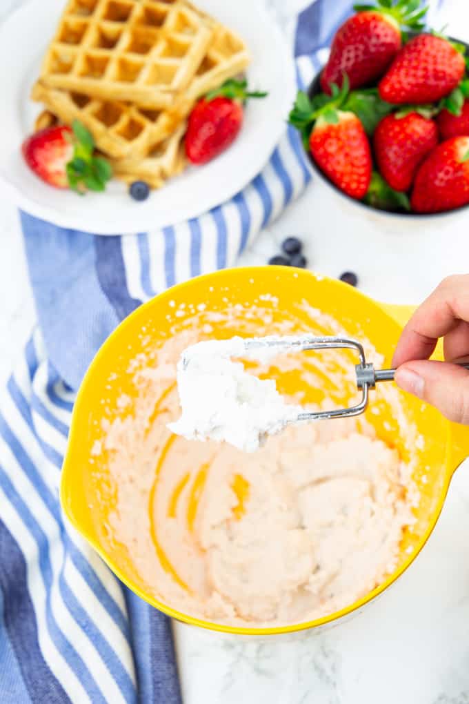 vegan whipped cream in a yellow bowl on a marble counter top with a hand holding a hand mixer with coconut whipped cream over the bowl