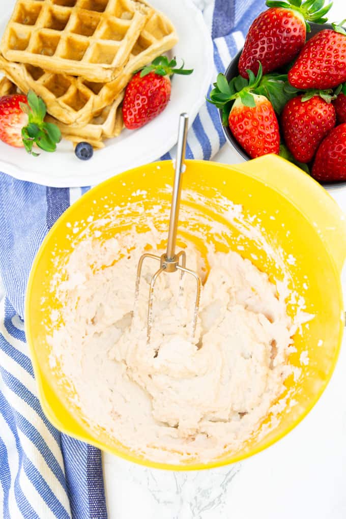 coconut whipped cream in a yellow mixing bowl on a marble counter top with a plate of waffles and strawberries in the background
