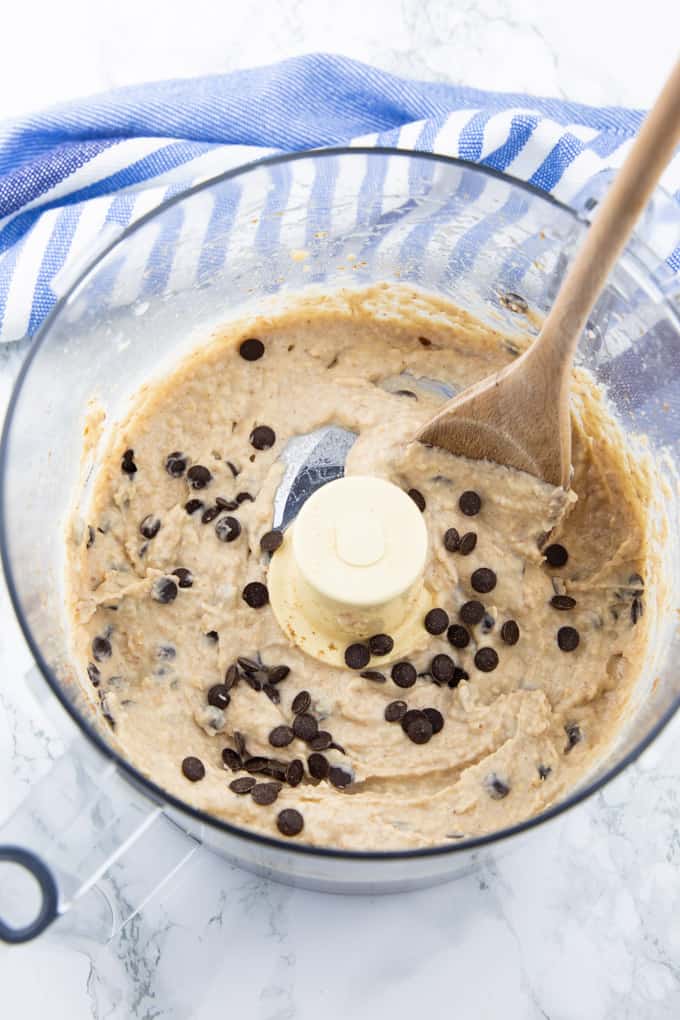 Vegan Cookie Dough with chocolate chips in a food processor with a wooden spoon on a marble counter top 