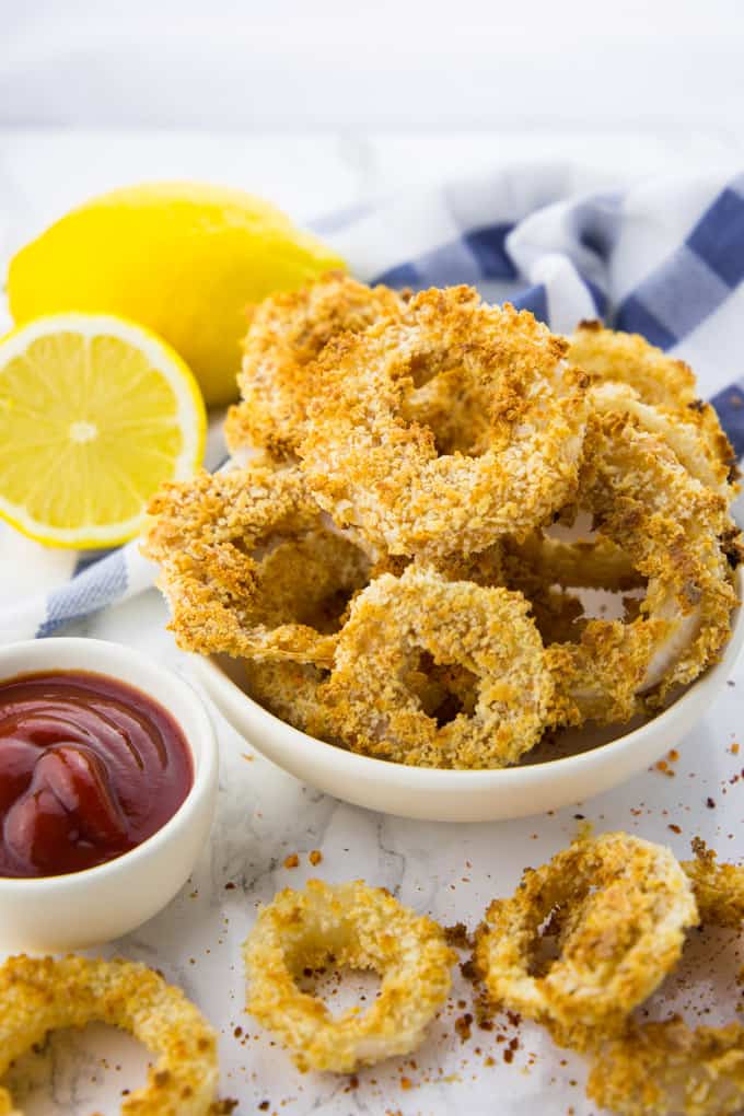 Vegan Onion Rings in a white bowl on a marble counter top with a small bowl of ketchup and more onion rings on the side 