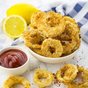 Vegan Onion Rings in a white bowl on a marble counter top with a small bowl of ketchup and more onion rings on the side