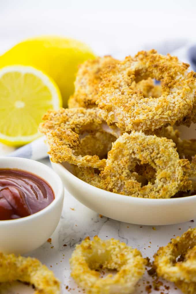 Vegan Onion Rings in a white bowl on a marble counter top with a small bowl of ketchup and lemons in the background 