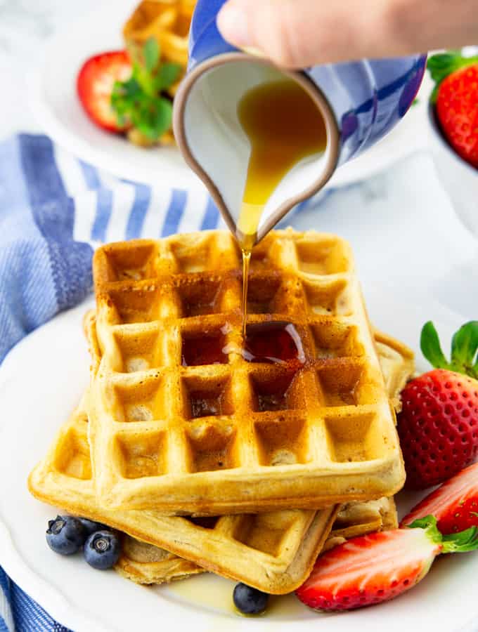 a stack of vegan waffles on a plate with one hand pouring maple syrup over it