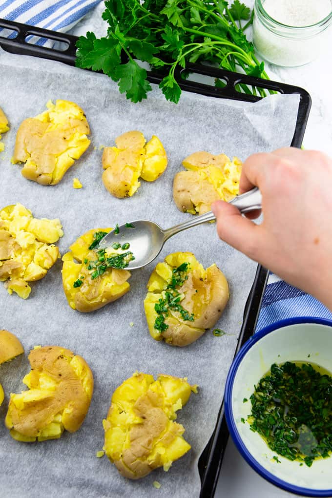 a parsley, olive oil, and garlic mixture is drizzled over smashed potatoes on a baking sheet lined with parchment paper with a bunch of parsley and a bowl of sea salt in the background