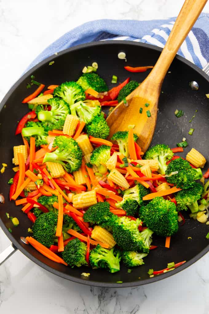vegetables for vegan Pad Thai in a large black pan with a wooden spoon on a marble countertop 