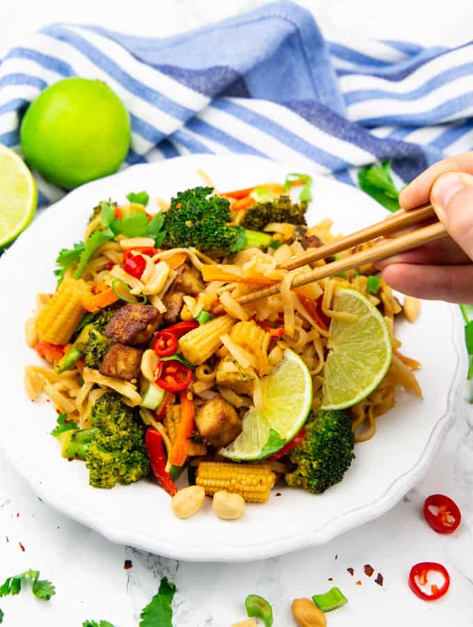 Vegan Pad Thai on a white plate with limes in the background and a hand picking up some noodles with chop sticks