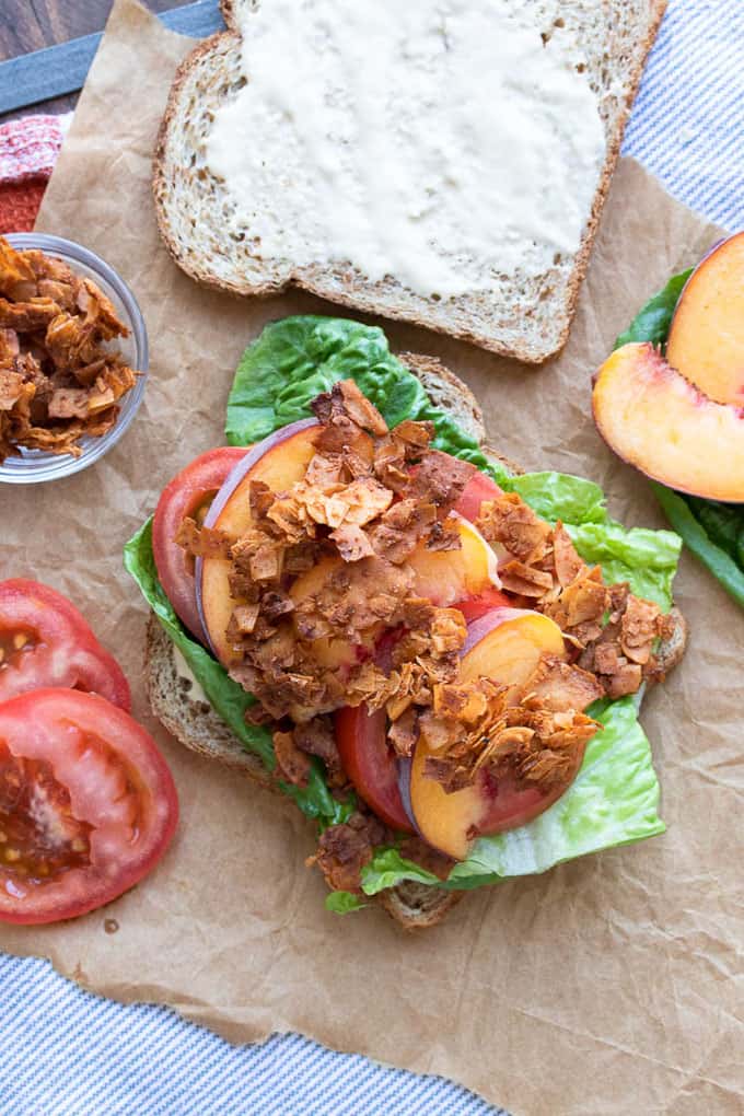 two vegan sandwiches with Vegan Coconut Bacon with Sliced Peaches on parchment paper with tomato slices and a bowl of vegan bacon on the side