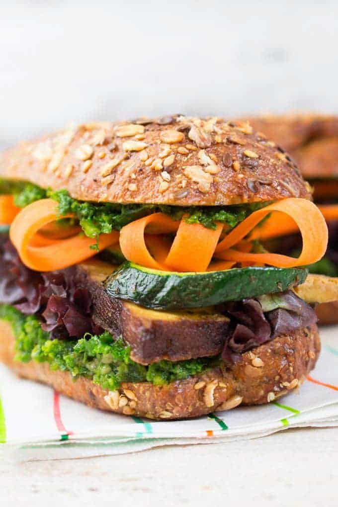 Two vegan sandwiches with smoked tofu, kale pesto, and carrots on a white wooden countertop with a striped napkin