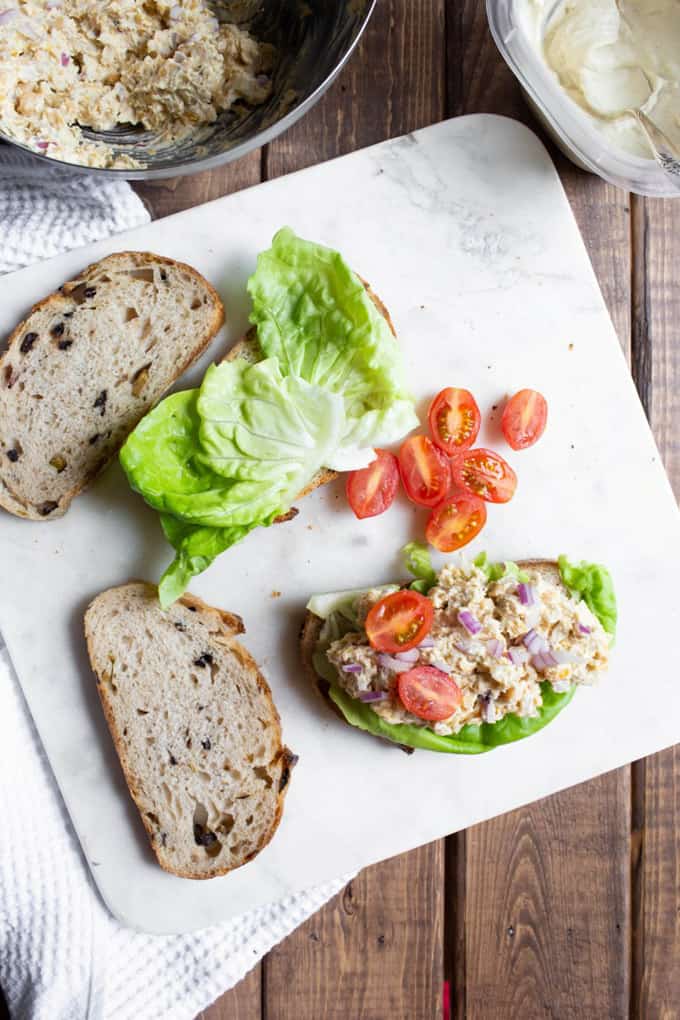 a vegan Caesar chickpea sandwich with two slices of bread, lettuce, and tomatoes on the side on a wooden countertop