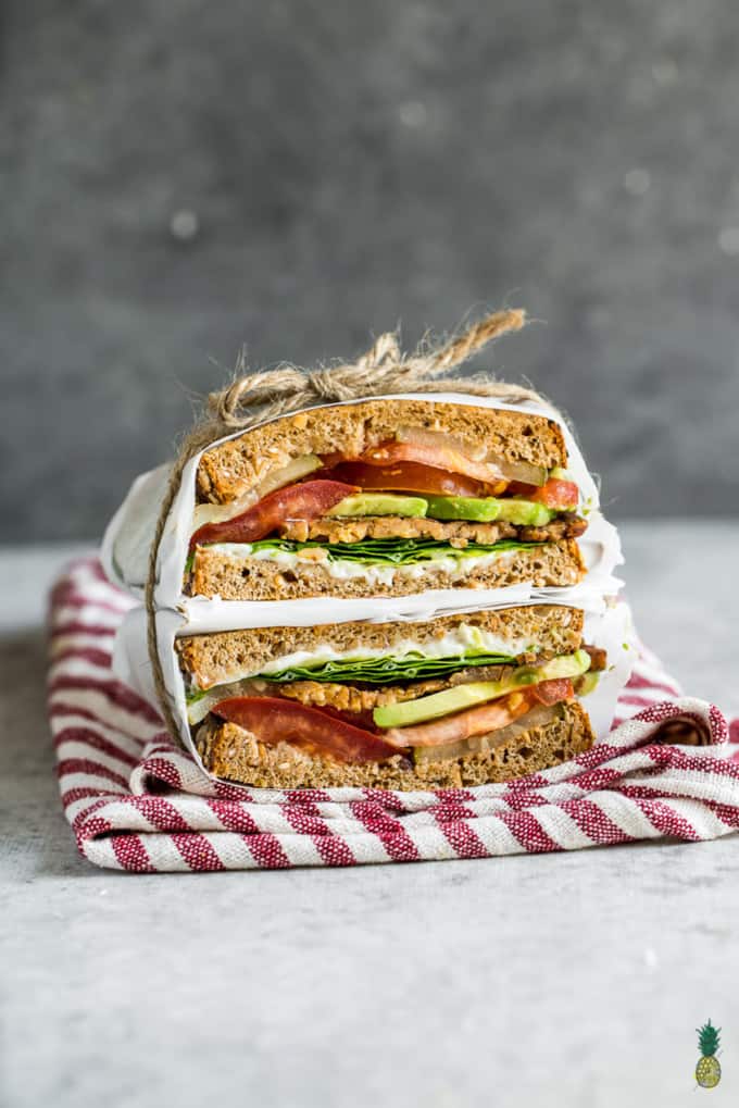 a stack of homemade vegan sandwiches with tempeh, lettuce, and tomato wrapped in parchment paper 