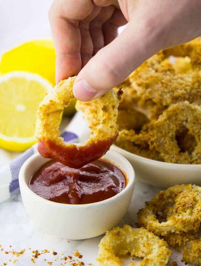 a small bowl with ketchup and a hand dipping a vegan onion ring into the bowl