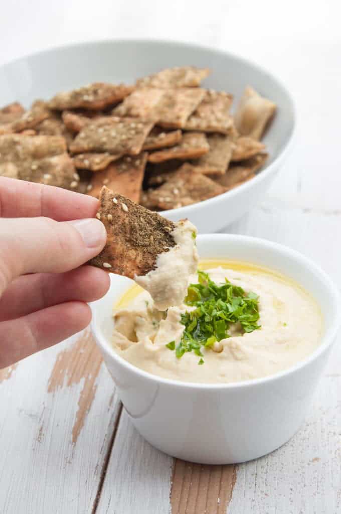 a hand dipping a Vegan Za’atar Cracker into a small bowl of hummus with more crackers in the background
