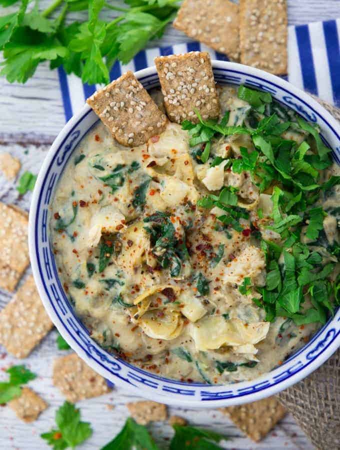 vegan spinach artichoke dip in a white and blue bowl with crackers and fresh parsley 