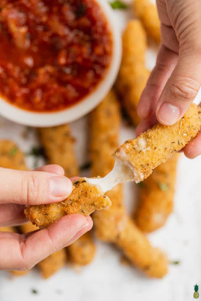 two hands holding a vegan mozzarella stick with a small bowl of marinara in the background